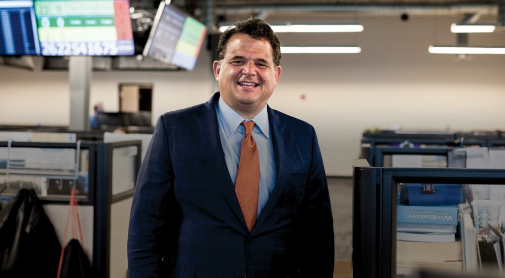 A photograph of a man with short brown hair in a deep blue suit with an orange tie smiling in an office setting.